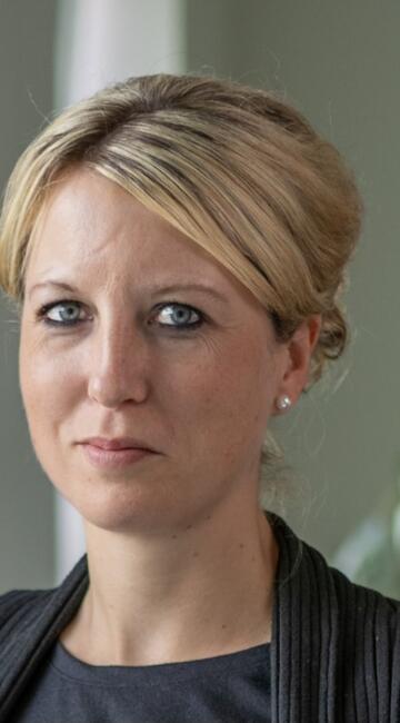 Eine Frau in einem Business-Outfit steht in einem Büroraum und schaut in die Kamera.
