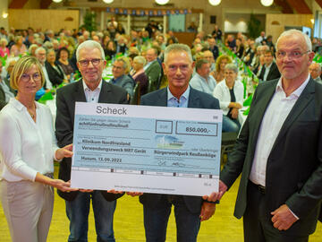 Vier Menschen halten einen großen Scheck mit der Summe 850.000 Euro in die Kamera. Im Hintergrund vollbesetzte Tische.