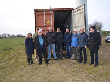 Mehrere Personen posieren draußen vor einem Container für ein Gruppenfoto. Im Hintergrund Felder und bewölkter Himmel.