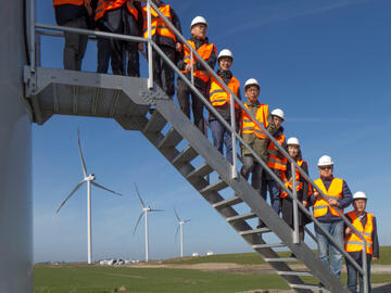 Koreanische Besucher in Warnwesten und Schutzhelmen stehen draußen auf einer Metalltreppe, im Hintergrund blauer Himmel.