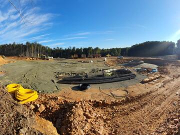 Panorama-Aufnahme einer sandigen Baustelle in einem Wald mit dem Fundament eines Windrads und Arbeitsgeräten ringsum.