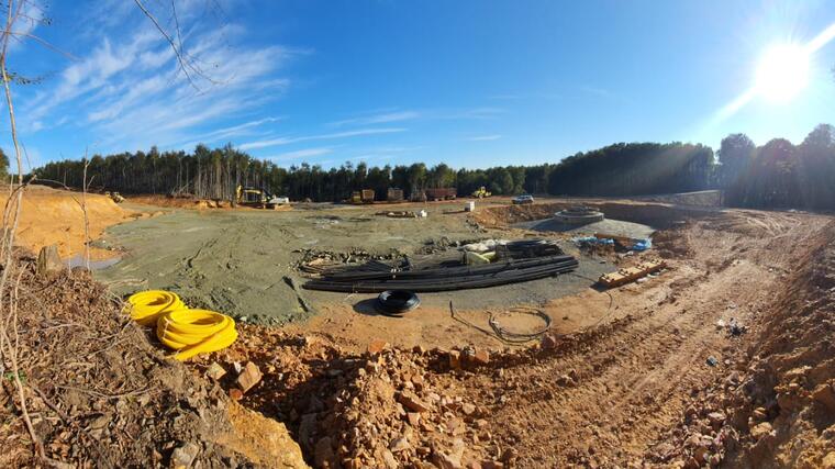 Panorama-Aufnahme einer sandigen Baustelle in einem Wald mit dem Fundament eines Windrads und Arbeitsgeräten ringsum.