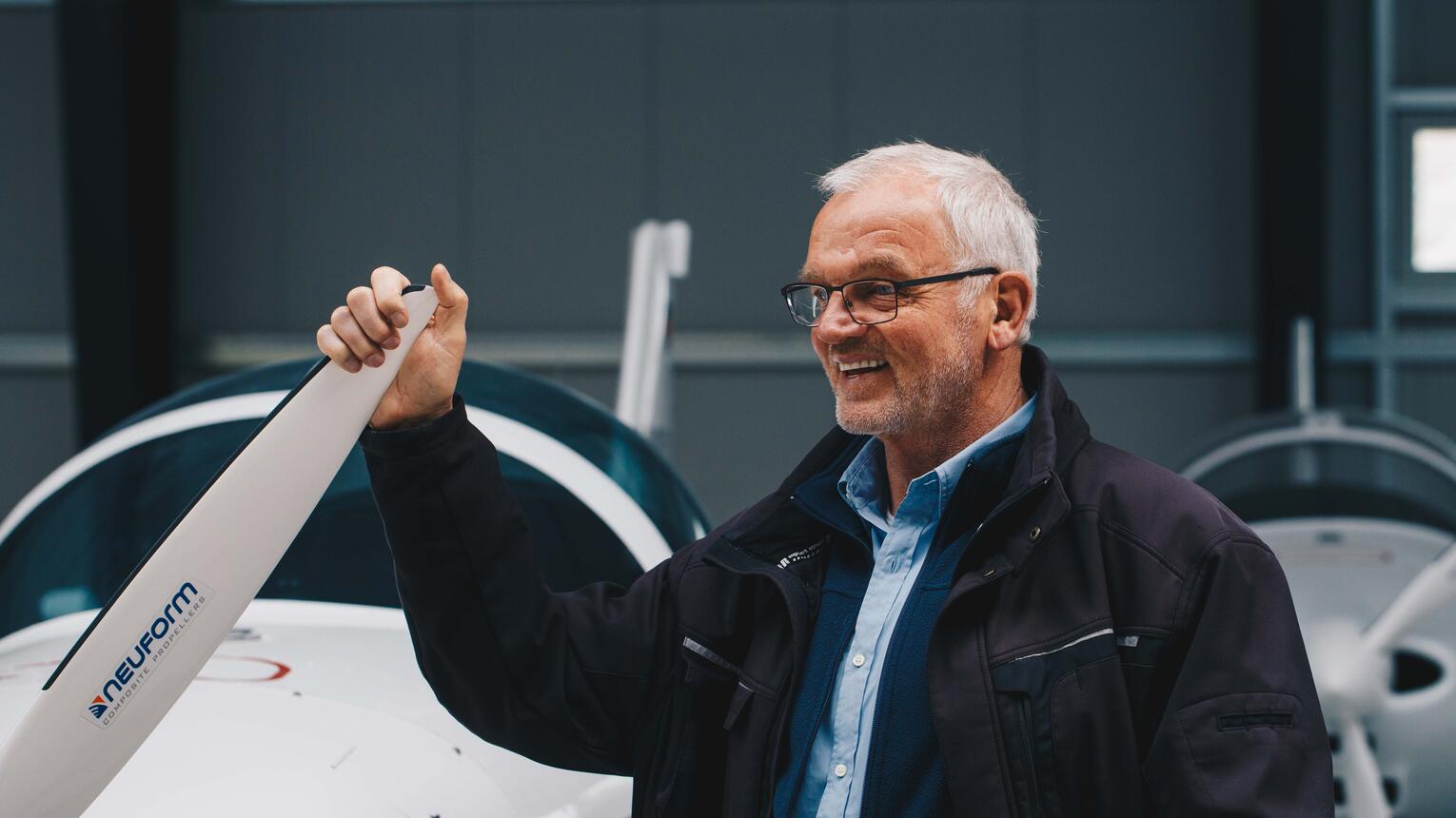 Geschäftsführer Dirk Ketelsen in der halbnahen Kameraeinstellung. Er steht in einem fabrikartigen Innenraum und hält ein schmales Bauteil oder einen Hebel fest.
