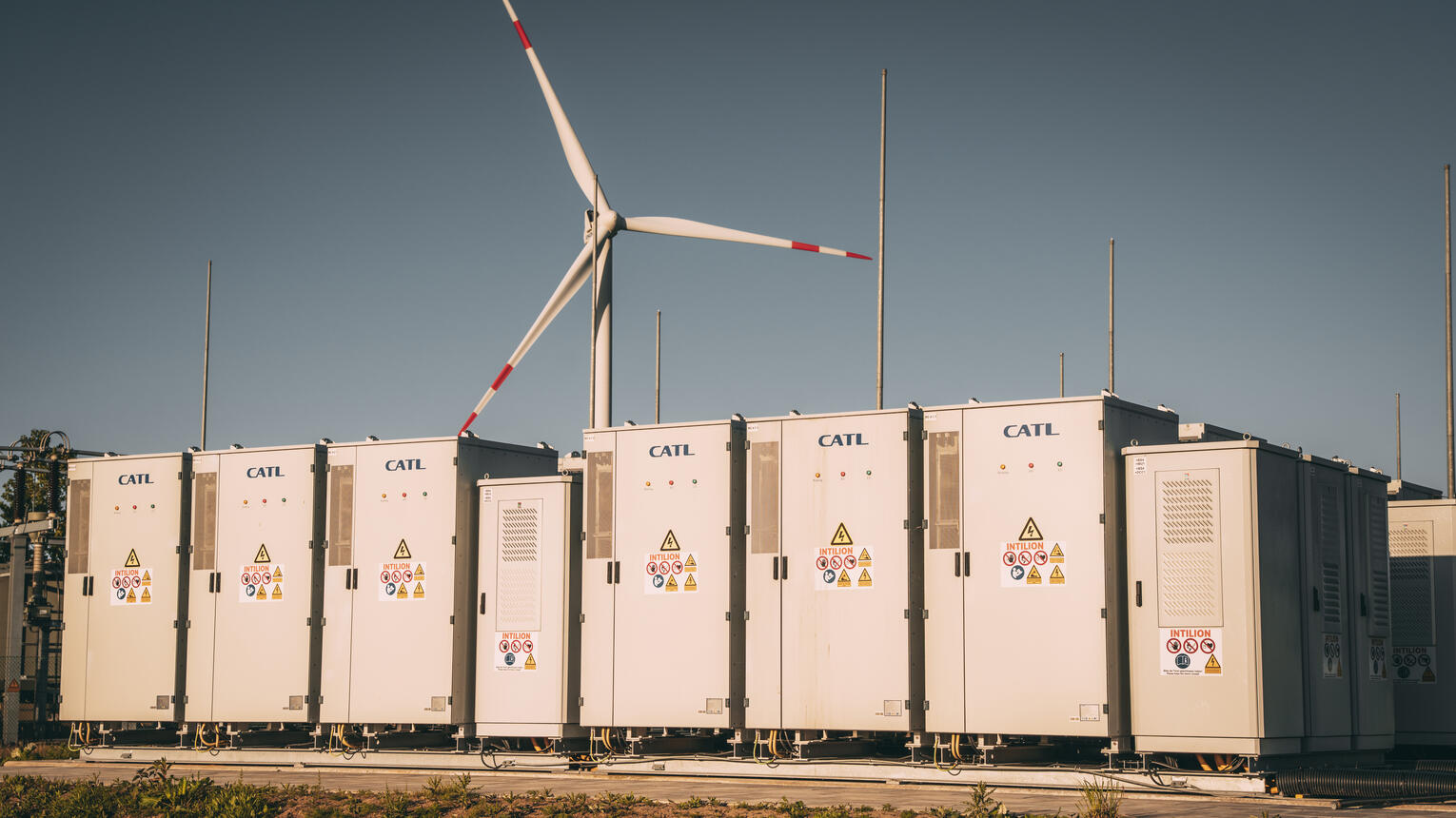 Das Umspannwerk eines Windparks, bei Sonnenschein an einer Straße. Im Hintergrund ein Windrad.
