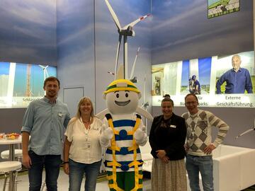 Am Dirkshof-Stand auf der WindEnergy-Messe posieren vier Mitarbeitende des Dirkshofs mit einem Maskottchen in Schutzausrüstung für ein Gruppenfoto.