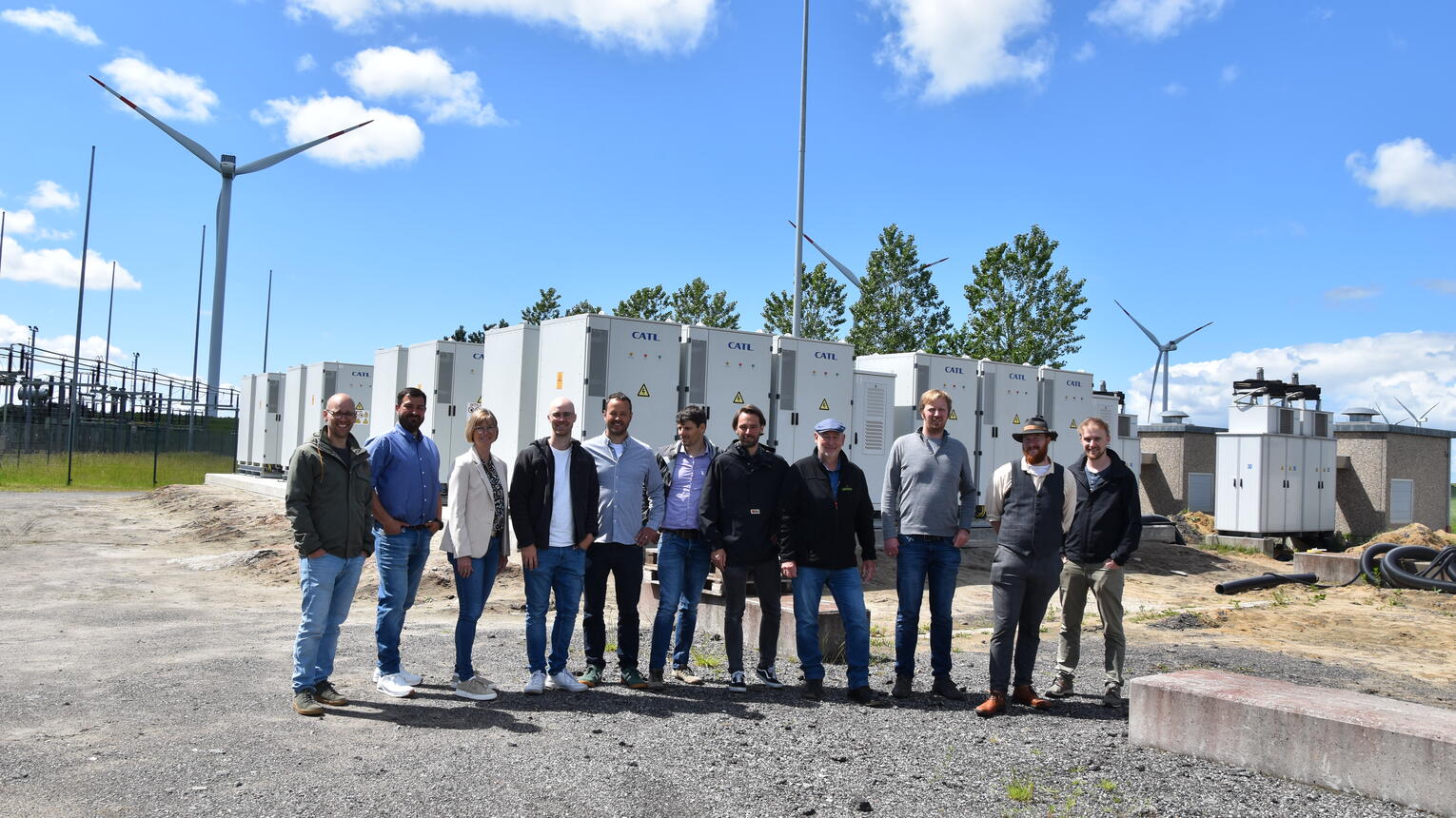 Eine Gruppe von lächelnden Menschen posiert vorm Umspannwerk für ein Foto. Im Hintergrund blauer Himmel.