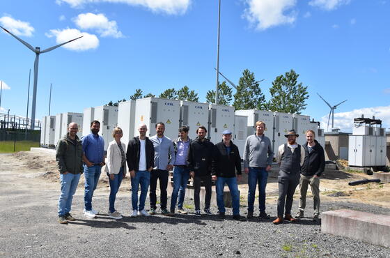 Eine Gruppe von lächelnden Menschen posiert vorm Umspannwerk für ein Foto. Im Hintergrund blauer Himmel.