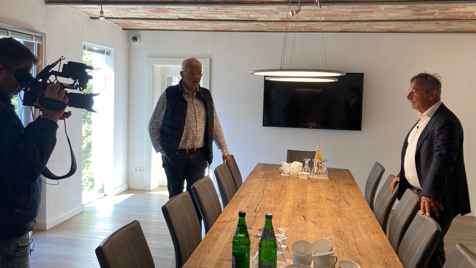 Geschäftsführer Dirk Ketelsen steht in einem Innenraum an einem Konferenztisch, ein anderer Mann steht ihm gegenüber. Ein Mann filmt.