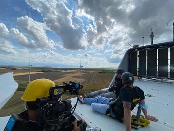 Ein Kameramann filmt, wie zwei Männer mit Schutzhelmen und Sicherungsharnischen entspannt oben auf einer Windturbine sitzen und übers Land schauen.