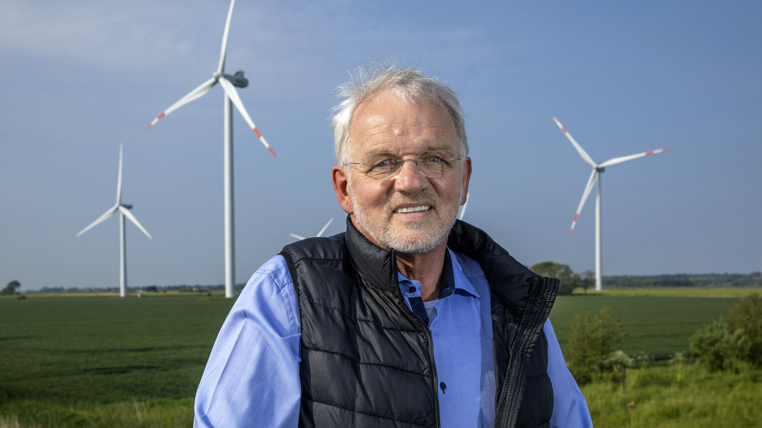 Geschäftsführer Dirk Ketelsen in einem blauen Hemd und einer schwarzen, offenen Weste, mit Windrädern und grünen Feldern im Hintergrund.