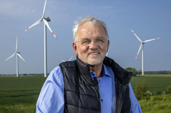 Geschäftsführer Dirk Ketelsen in einem blauen Hemd und einer schwarzen, offenen Weste, mit Windrädern und grünen Feldern im Hintergrund.