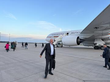 Dirk Ketelsen steht lachend am Flughafen von Ankara vor einem Flugzeug, im Hintergrund blauer Himmel.