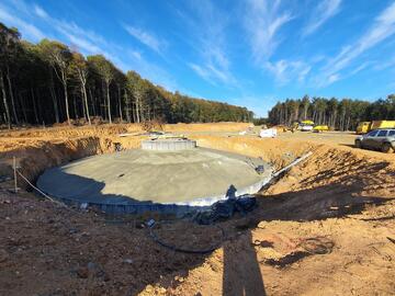 Eine sandige Baustelle in einem Wald mit dem Fundament eines Windrads und Arbeitsgeräten ringsum. Schatten fallen auf die Baustelle.