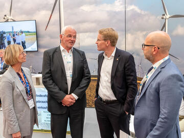 Der Ministerpräsident Daniel Günther, Dirk Ketelsen und zwei weitere Personen unterhalten sich bei der HusumWind vor einem großen Plakat mit Windrädern darauf.
