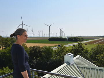 Nina Scheer, eine Frau mittleren Alters mit Brille, dunklem Haar und dunklem Longsleeveshirt, blickt bei Sonnenschein von einem Aussichtspunkt aus über grüne Felder, Bäume und Windräder.
