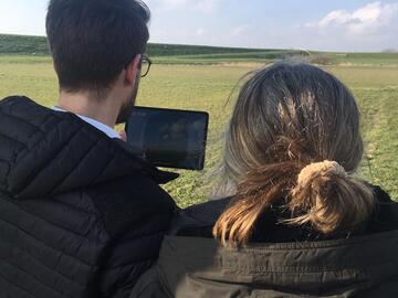 Blick über die Schulter eines Manns und einer Frau, die sich draußen auf einem Feld auf dem Tablet ein Video angucken.