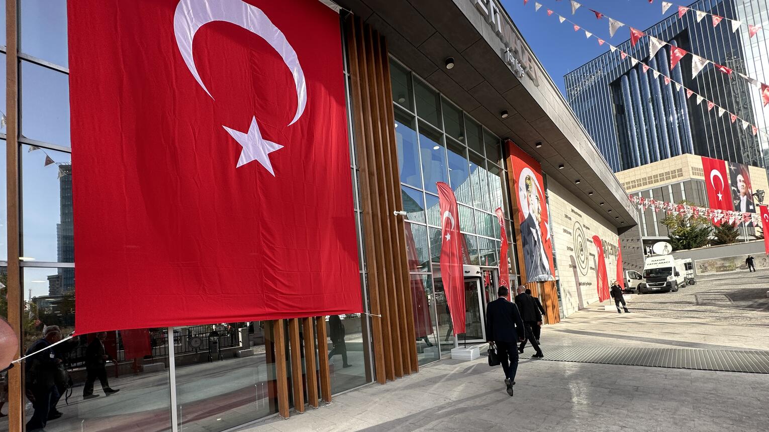 Ein verglastes Gebäude mit einer großen türkischen Flagge, die davon herunterhängt. Rot-weiße Wimpeln verzieren den Platz davor.