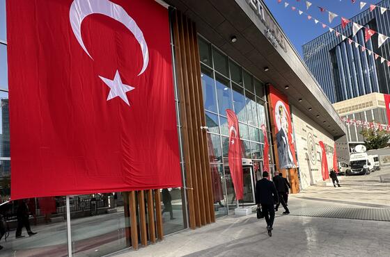 Ein verglastes Gebäude mit einer großen türkischen Flagge, die davon herunterhängt. Rot-weiße Wimpeln verzieren den Platz davor.