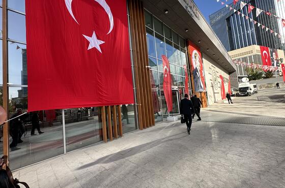 Ein verglastes Gebäude mit einer großen türkischen Flagge, die davon herunterhängt. Rot-weiße Wimpeln verzieren den Platz davor.