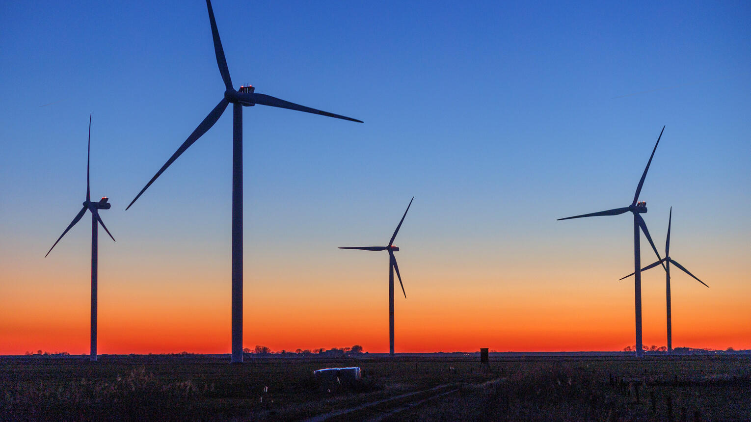 Dunkle Silhouetten von Windrädern vor einem orange-blauen Abendhimmel. An den Turbinen der Windräder blinkt es rot.